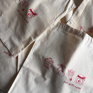 Op de afbeelding: Crèmekleurige canvas draagtassen met rode illustraties van kersen, bloemen en jampotten. De zin "fresh off the market" is in het rood gedrukt. De tassen hebben een eenvoudig, praktisch ontwerp.