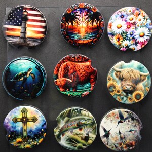 May include: Nine round car coasters with various designs. The designs include a lighthouse with an American flag, a sunset over the ocean, a field of daisies, a sea turtle, a Highland cow, a cross, a fish, and hummingbirds.