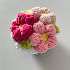 Può includere: Una composizione floreale in peluche fatta a mano con un gruppo di fiori morbidi e strutturati. I fiori sono in tonalità di rosa, fucsia e pesca, con centri gialli e foglie verdi. La base è bianca.
