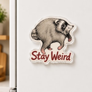 May include: A clear acrylic refrigerator magnet featuring a detailed black and white illustration of a round, furry raccoon-like creature with pinkish paws. Below the animal, the text Stay Weird is printed in a bold, dark red sans-serif font.