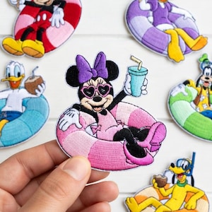 Puede incluir: Parches bordados con los personajes de Disney Mickey Mouse, Minnie Mouse, Donald Duck, Daisy Duck, Goofy y Pluto. Cada personaje está representado relajándose en un flotador inflable colorido, sosteniendo una bebida o un helado. Los parches están sobre una superficie de madera blanca.