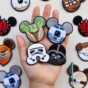 Op de afbeelding: Een verzameling geborduurde patches met Star Wars-personages in een Mickey Mouse-oordesign. Personages zijn onder meer Darth Vader, Yoda en R2-D2. De patches zijn kleurrijk en gedetailleerd, met zwarte, witte, bruine en blauwe accenten.