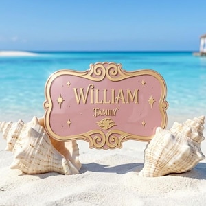 Op de afbeelding: Een rechthoekig roze bord met een sierlijke gouden rand met de tekst "WILLIAM FAMILY" in gouden letters, geaccentueerd door kleine gouden sterren en een centraal gouden embleem, geplaatst tussen twee grote, lichtgekleurde schelpen.