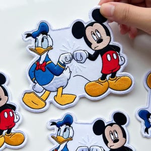 May include: Embroidered patches featuring Mickey Mouse and Donald Duck characters. Mickey Mouse is in red shorts and yellow shoes, while Donald Duck wears a blue sailor suit. The patches have white borders and are on a white surface.