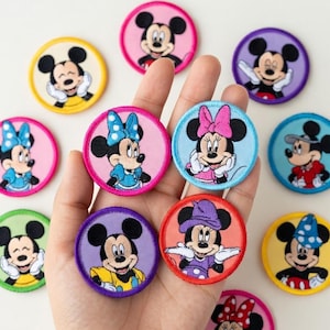 May include: A selection of circular embroidered patches showcasing Mickey and Minnie Mouse in diverse poses and attire. The patches feature a range of colours, including pink, blue, yellow, and purple. The patches are held in a hand against a white backdrop.