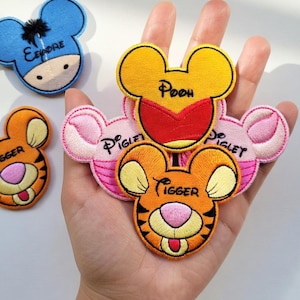 May include: Embroidered patches of Winnie the Pooh characters, including Tigger, Piglet, Pooh, and Eeyore. The patches are shaped like Mickey Mouse ears. Tigger is orange and black, Piglet is pink, Pooh is yellow, and Eeyore is blue.