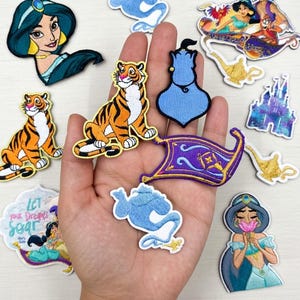 Puede incluir: Una colección de parches bordados con personajes y elementos de la película animada Aladdin. Los parches incluyen a Jasmín, el Genio, Abu, una alfombra mágica, un castillo y un tigre. Los parches son coloridos y detallados, con varias formas y tamaños.