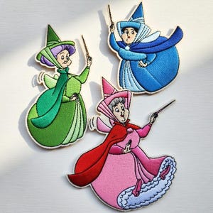 May include: Three embroidered patches featuring cartoon fairy characters in green, blue, and pink dresses with matching hats and capes. Each fairy holds a wand. The patches have a white border and are set against a light background.