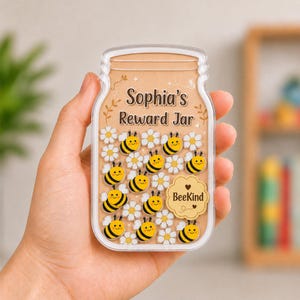 May include: A clear acrylic jar-shaped reward board featuring the text Sophia's Reward Jar at the top. The interior is filled with small, colorful bee and white daisy icons. A scalloped badge in the center displays the text BeeKind with a heart.