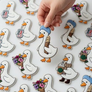 Puede incluir: Parches bordados con patos blancos con picos y patas naranjas. Algunos patos llevan boinas azules, sostienen flores, beben de latas de Diet Coke o llevan baguettes. Los parches tienen un borde blanco y están sobre una superficie blanca.