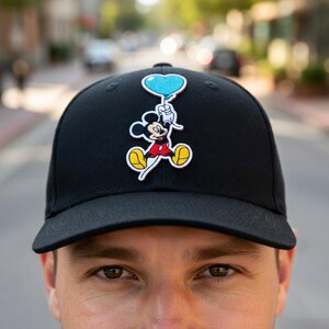 May include: Black baseball cap featuring a Mickey Mouse patch holding a blue heart-shaped balloon. The cartoon character is wearing a red shirt, yellow shoes, and white gloves. The cap is a classic six-panel design.