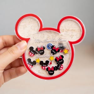 Op de afbeelding: Een helder, Mickey Mouse-vormig frame met een rode omtrek. Binnenin bevinden zich verschillende zwarte Mickey Mouse-hoofden met rode strikken en het woord "Name". De tekst "Mrs. Smith's Class 2025-2026 Back to School" is ook zichtbaar.