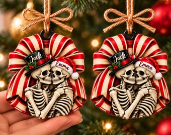 Könnte beinhalten: Zwei identische flache Weihnachtsschmuck-Anhänger mit einem Paar sich umarmender Skelette. Ein Skelett trägt einen schwarzen Zylinder mit der Aufschrift Jack und Stechpalme, das andere eine rote Weihnachtsmütze mit der Aufschrift Emma, umrahmt von einer rot-cremefarbenen Schleife.