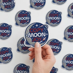 May include: Embroidered patch with a celestial design. The patch features a blue background with white stars, a crescent moon, and a rocket ship. The text "COPY MOON JOY" is embroidered in red and white. Several patches are visible.