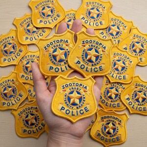 May include: Embroidered yellow Zootopia Police badge patches with the words "Zootopia Police" and "Integrity, Trust, Bravery" around a star emblem. Several patches are visible, some held in a hand.