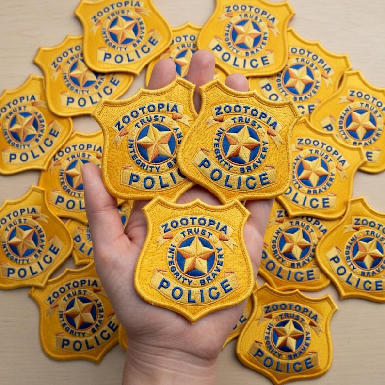 May include: Embroidered yellow Zootopia Police badge patches with the words "Zootopia Police" and "Integrity, Trust, Bravery" around a star emblem. Several patches are visible, some held in a hand.
