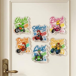 Peut inclure: Un ensemble de cinq autocollants muraux en acrylique repr&eacute;sentant les personnages Nintendo Yoshi, Mario, Peach, Daisy, Luigi et Bowser dans des v&eacute;hicules de course. Chaque autocollant inclut le nom du personnage, l'ann&eacute;e 1992 et des motifs &agrave; damier.