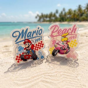 Peut inclure: Deux pr&eacute;sentoirs en acrylique transparent repr&eacute;sentant Mario dans un kart rouge et Peach sur une moto rose. Chaque pi&egrave;ce affiche le nom du personnage en cursive, l'ann&eacute;e 1992 et un motif &agrave; damier. Le mod&egrave;le Mario utilise du bleu et du rouge, tandis que celui de Peach utilise des tons rose et jaune.