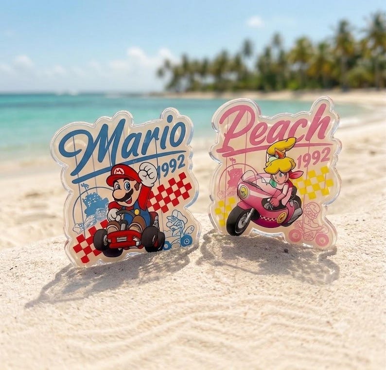 Peut inclure: Deux pr&eacute;sentoirs en acrylique transparent repr&eacute;sentant Mario dans un kart rouge et Peach sur une moto rose. Chaque pi&egrave;ce affiche le nom du personnage en cursive, l'ann&eacute;e 1992 et un motif &agrave; damier. Le mod&egrave;le Mario utilise du bleu et du rouge, tandis que celui de Peach utilise des tons rose et jaune.