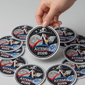 May include: Circular embroidered patches with a black background, featuring the text "ARTEMIS 2026", a rocket, the moon, and Earth. The patches have a white border and are being held by a hand. The patches are likely for a space mission.