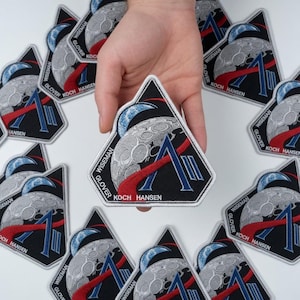 May include: A collection of embroidered patches with a triangular design. The patches feature a stylized moon, a blue and red graphic, and the names WISEMAN, GLOVER, KOCH, and HANSEN. The patches are black with white and blue accents.
