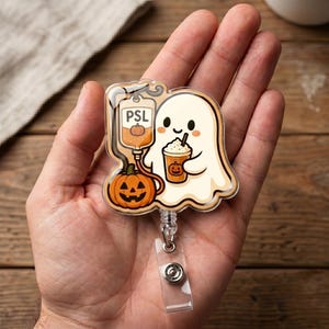 May include: An acrylic badge reel featuring a cartoon ghost holding a pumpkin spice latte, connected to an IV bag labeled PSL with a small pumpkin icon. The design includes a bright orange jack-o'-lantern at the base, all set on a clear backing.