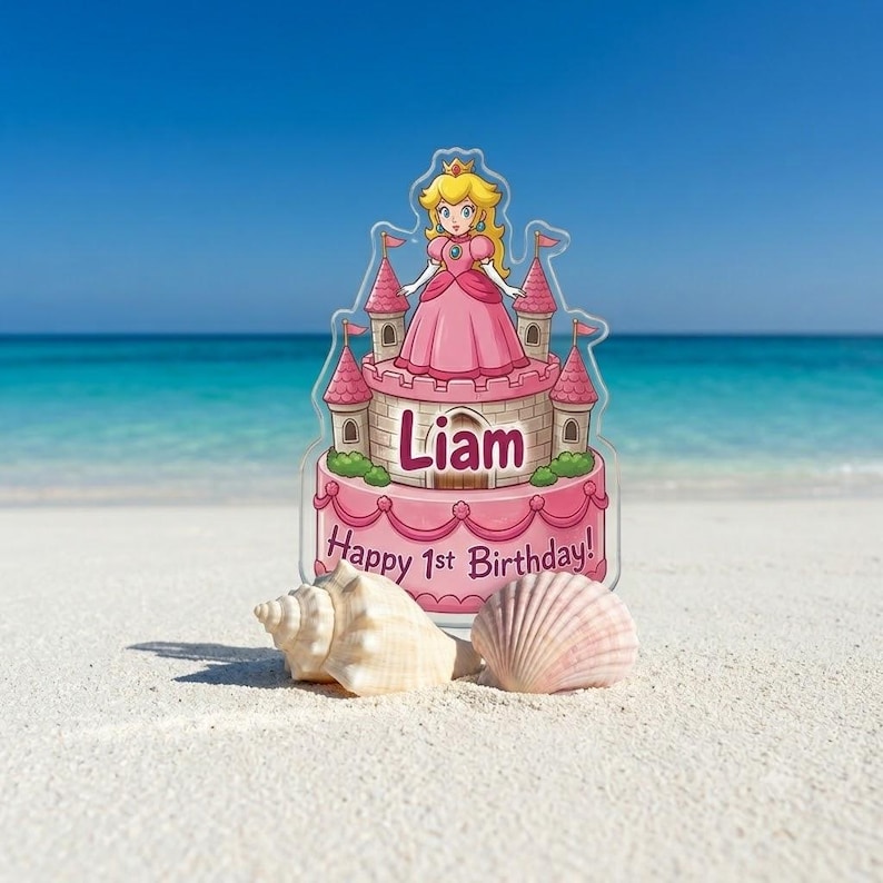 May include: A clear acrylic cake topper featuring a cartoon princess in a pink dress standing atop a pink castle. The castle base displays the name Liam and the text Happy 1st Birthday. Two seashells are positioned in front of the topper.