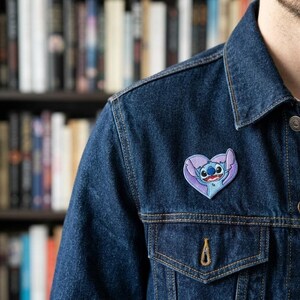 Może przedstawiać: Granatowa kurtka dżinsowa z naszywką w kształcie serca przedstawiającą postać z kresk&oacute;wki. Naszywka ma niebiesko-fioletową kolorystykę. Kurtka ma klasyczny kr&oacute;j z widocznymi szwami i zapięciem na guziki.