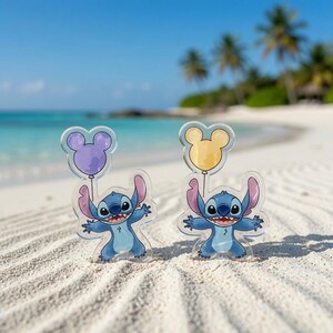 May include: Two clear acrylic standees featuring the blue alien character Stitch, each holding a balloon shaped like a mouse head. One balloon is light purple and the other is light yellow. The figures have large ears, wide smiles, and dark eyes.