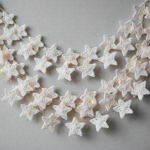 May include: A string of white, crocheted star-shaped lights. The stars are evenly spaced along a thin, clear wire. The lights are illuminated, casting a soft glow. The background is a plain, light gray surface. The overall theme is festive and decorative.
