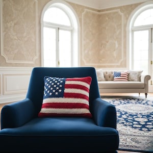 Pode incluir: Uma almofada de crochê com o design da bandeira americana, com um campo azul de estrelas brancas e listras vermelhas e brancas. A almofada está colocada em uma poltrona azul em um quarto bem iluminado.