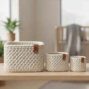 Può includere: Tre cesti portaoggetti intrecciati color crema di diverse dimensioni sono esposti su una superficie di legno. Ogni cesto ha una trama intrecciata e una piccola etichetta in pelle marrone con la lettera "H". I cesti sono disposti dal più grande al più piccolo.
