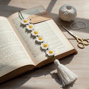 Pode incluir: Um marcador de corrente de margaridas feito à mão com flores de crochê brancas e amarelas. O marcador está colocado em um livro aberto com uma borla na extremidade. Uma bola de fio branco e tesouras douradas estão ao lado do livro. O marcador tem uma etiqueta que diz "Daisy Chain Bookmark".