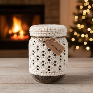 May include: A clear glass jar filled with coffee beans, wrapped in a cream-colored, openwork crochet cozy. A small brown tag with the word "Handmade" hangs from a twine bow around the jar. The background features a fireplace and a Christmas tree.