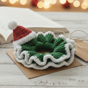 Puede incluir: Una scrunchie hecha a mano con temática navideña. La scrunchie es verde con ribete blanco y un pequeño gorro de Papá Noel rojo y blanco. La scrunchie está hecha de hilo y se encuentra sobre una tarjeta marrón.