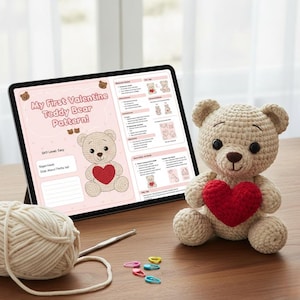 Puede incluir: Un oso de peluche de ganchillo con un corazón rojo junto a una tableta que muestra un patrón de San Valentín. Una madeja de hilo, un ganchillo y marcadores de punto están sobre una mesa de madera. El patrón se titula "My First Valentine Teddy Bear Pattern!"