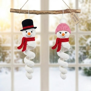 May include: Two handmade crochet snowman ornaments hanging from a wooden branch. One snowman wears a black top hat and red scarf, the other a pink hat and red scarf. Both have white spiral bodies and the tag reads "Handmade Pattern".