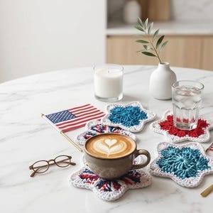 May include: A coffee cup with latte art sits on a star-shaped crochet coaster with red, white, and blue colors. Other coasters, a candle, a glass of water, and small American flags are on a marble table.