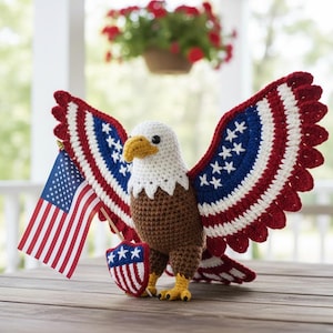 May include: A crocheted bald eagle with outstretched wings, holding a miniature American flag and a shield. The eagle features red, white, and blue wing patterns, a white head, brown body, and yellow beak and feet. The patriotic craft is displayed on a wooden surface.
