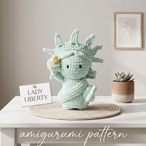May include: A crocheted Lady Liberty amigurumi in light blue, holding a yellow torch. The amigurumi is on a beige round mat, with a card that says "LADY LIBERTY". The words "amigurumi pattern" are written at the bottom.