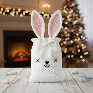 May include: A white crocheted bunny bag with pink inner ears and a drawstring closure. The bunny face is embroidered with black thread. The bag is sitting on a wooden surface with candies.