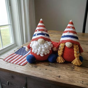 May include: Two handmade crochet gnomes with red, white, and blue patriotic colors. One gnome has a white beard, the other has blonde braids. Both have star-studded hats and blue feet. An American flag is placed nearby.