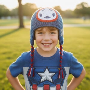 May include: A blue crocheted hat with a white star emblem and red and white accents. The hat has braided earflaps with red and white tassels. The child is wearing a blue t-shirt with a star graphic.