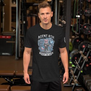 May include: A dark heather gray short-sleeve t-shirt featuring a graphic of a cat wearing a suit and sunglasses, labeled Agent Ash, with the text Drop The Mic below it. The design includes a microphone and abstract blue circular elements.