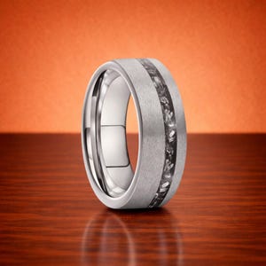 May include: A silver-colored ring with a textured, matte finish. The ring features a central inlay of dark, speckled material. The ring is set against a warm, orange background and a wooden surface.