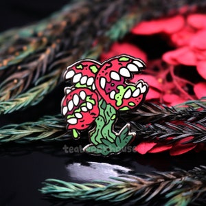 May include: A red and green enamel pin featuring a cartoon plant with three mouths. The plant has a green stem and leaves. The mouths are open and have sharp teeth.