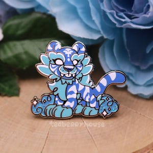 以下が含まれることがあります： 白と青のパターンでデザインされた、漫画の虎の青と白のエナメルピン。虎はキラキラのアクセントのある雲の上に座っています。