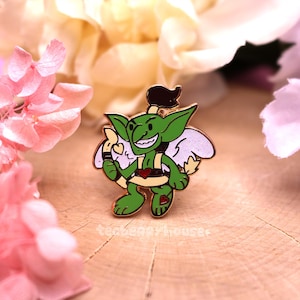 Puede incluir: Un pin de esmalte de cupido goblin verde con un corazón rojo, alas blancas y un arco y flecha dorados. El goblin lleva un cinturón marrón y tiene una expresión traviesa.