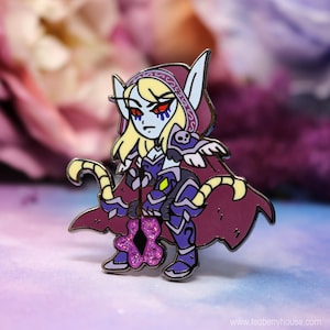 May include: A purple and blue enamel pin of a female elf character with a purple hooded cloak, holding a purple crystal staff. The character has blue eyes, blonde hair, and a dark blue and purple outfit.