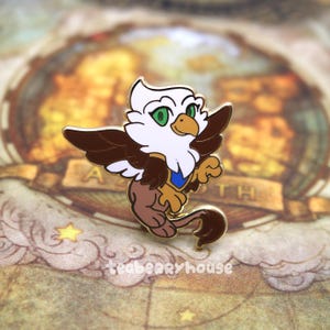 May include: A gold-colored enamel pin of a white and brown griffin with blue accents. The griffin has green eyes and a brown tail.